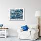 Vathi – atmospheric abstract painting with layered blue, white, and subtle gold textures, framed in light wood, styled above a white armchair with blue cushions in a coastal-inspired living room.